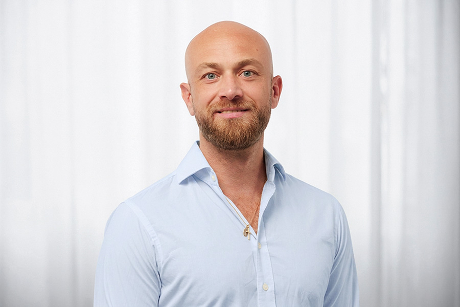 Smiling man with a shaved head and beard wearing a light blue button-up shirt, standing against a white curtain background (head-and-shoulders portrait). Gently lit indoor portrait.