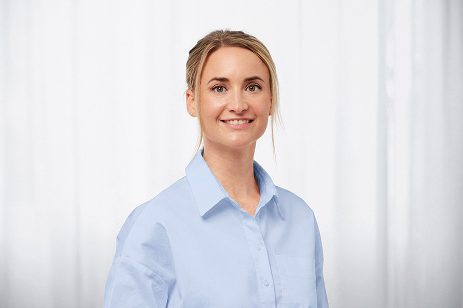 Smiling woman in a light blue button-down shirt poses against a pale backdrop, a professional headshot.