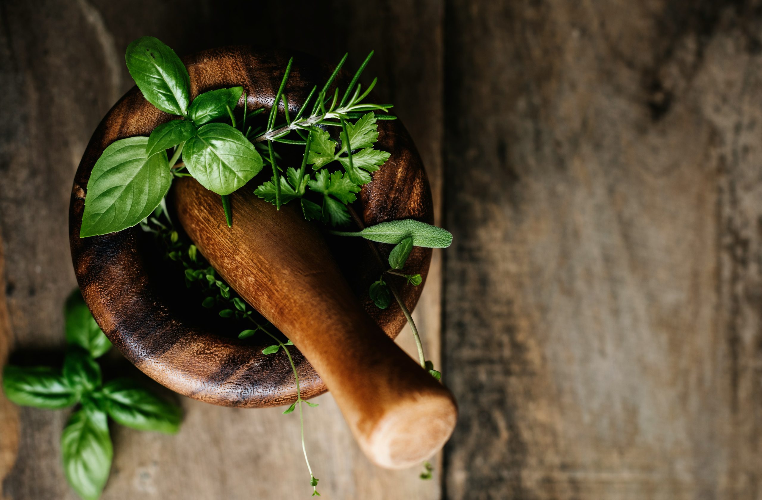 Fresh green herbs (basil, rosemary, parsley) resting in a dark wooden mortar on a rustic wooden surface.