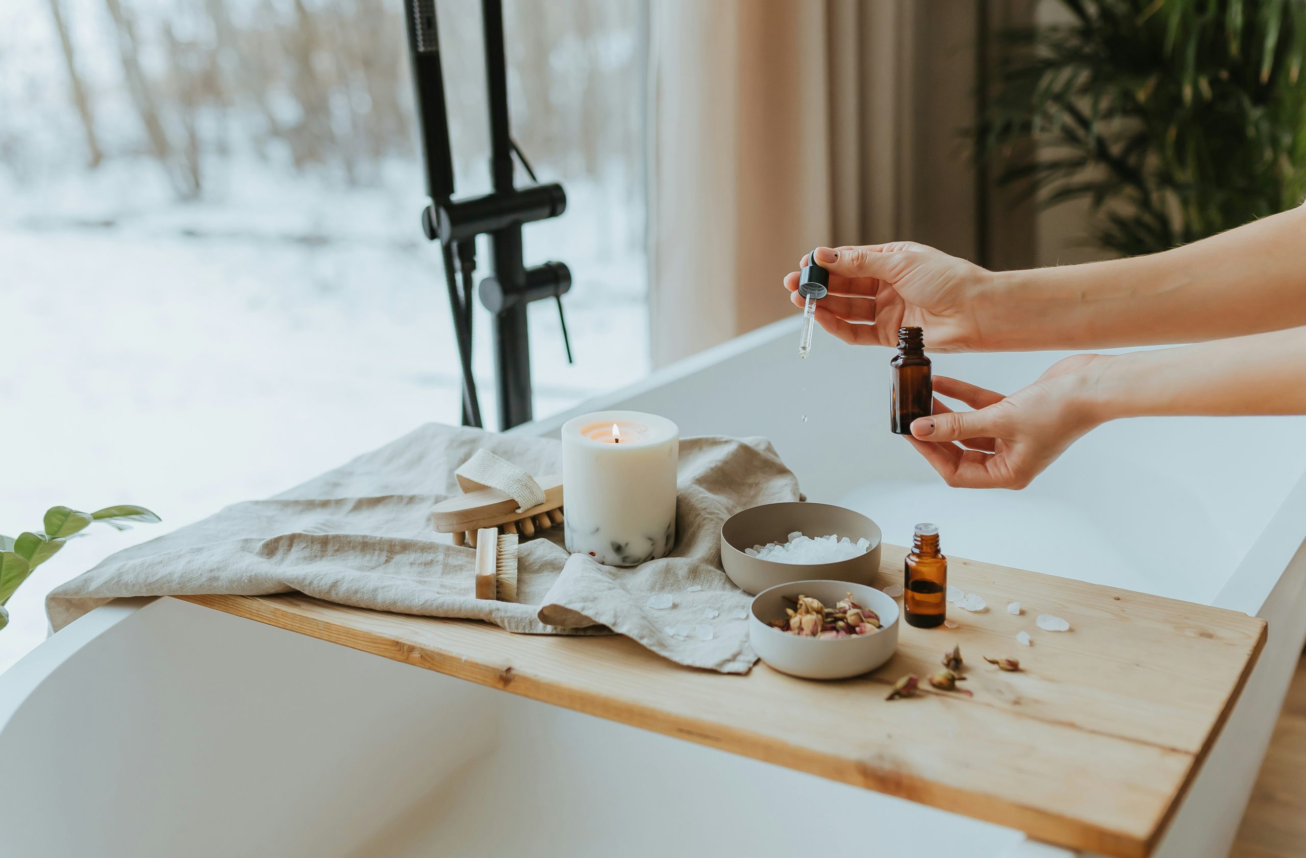 Hands holding a dropper over a brown essential oil bottle on a wooden table with a lit candle and bowls nearby in a spa setup.