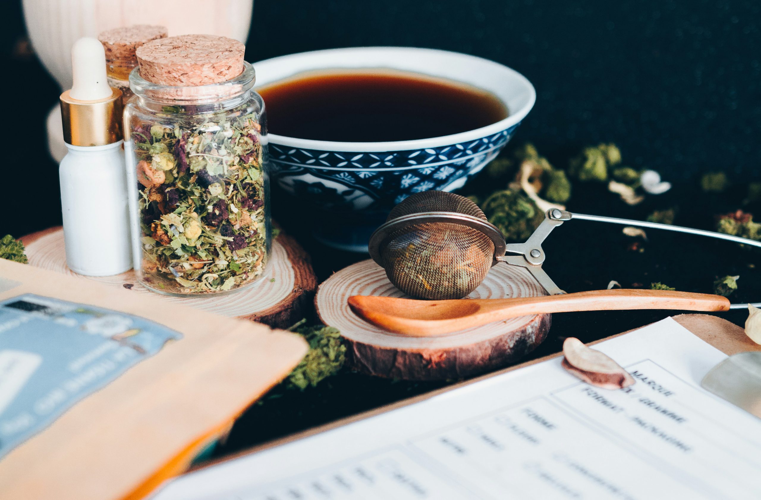 Jar of dried herbal tea leaves with cork lid beside a blue patterned teacup and metal infuser on a wooden board, ready for brewing